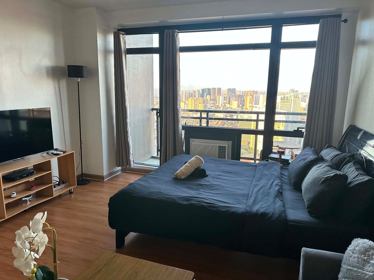 Queen bed and sofa beside floor-to-ceiling balcony windows in daylight.